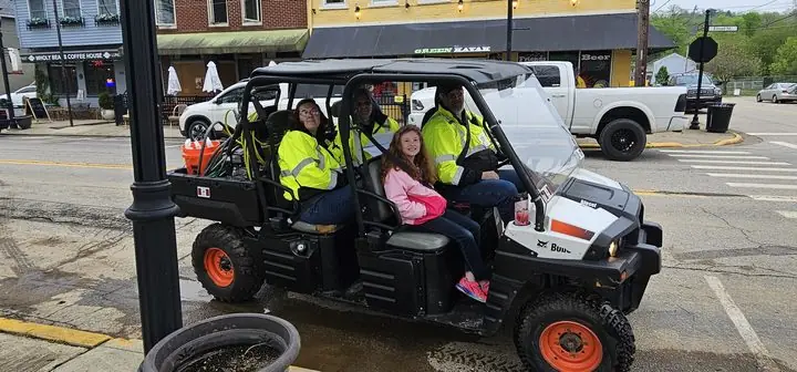 Volunteers in high-visibility jackets ride a utility vehicle down a small-town main street