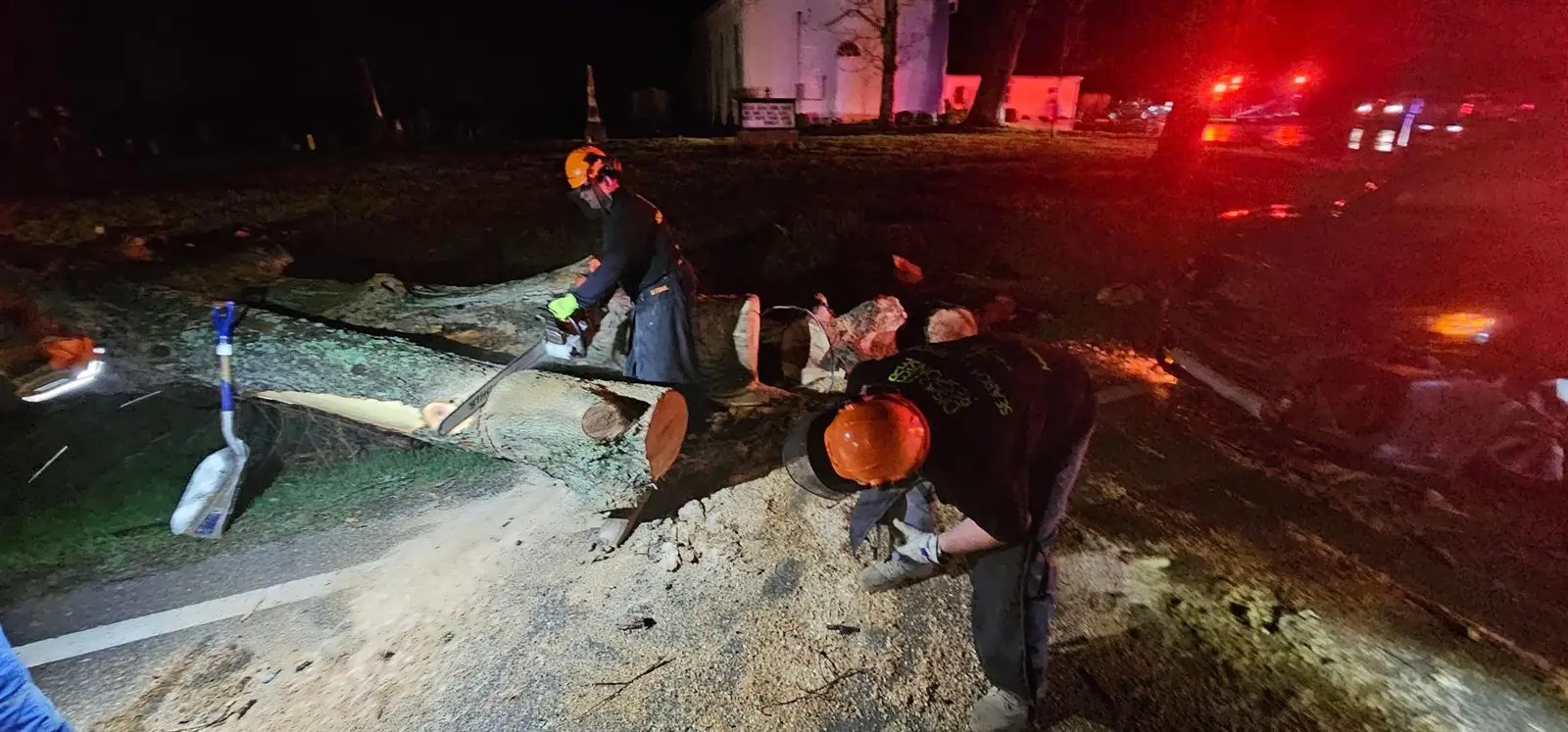 American Regulators USA volunteers in helmets cut a fallen tree off the road at night as emergency lights flash nearby